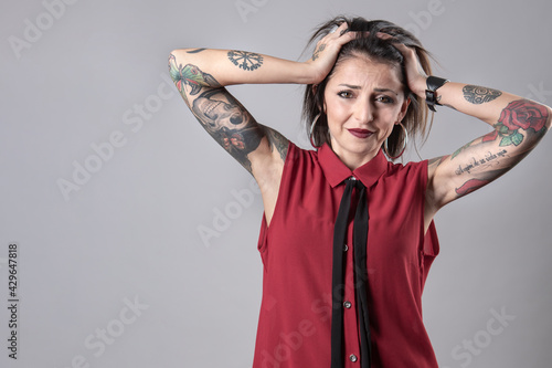 ragazza  mora con i tatuaggi indossa una camicia senza maniche rossa, porta le mani alla testa in segno di disperazione,  isolata su sfondo grigio