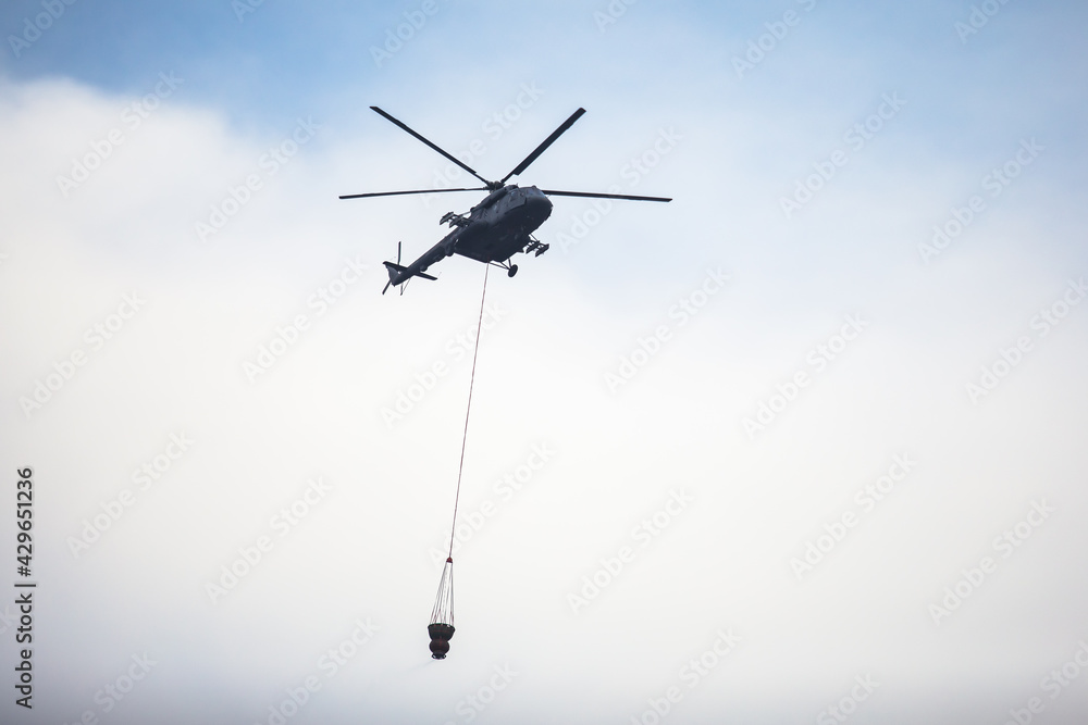 Fototapeta premium Fire fighting helicopter silhouette with bambi bucket for сarrying water to put out a massive building city fire, process of put out a large blaze bush fire wildifre, aerial firefighting with chopper