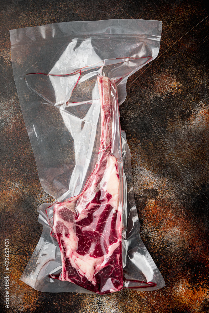 Tomahawk steak vacuum packed dry aged beef marbled meat for sous vide, on old dark rustic background, top view flat lay