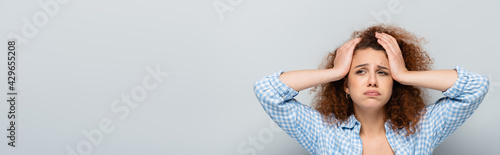exhausted woman with curly hair holding hands on head on grey background, banner