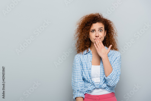 embarrassed woman covering mouth with hand while looking at camera on grey background photo