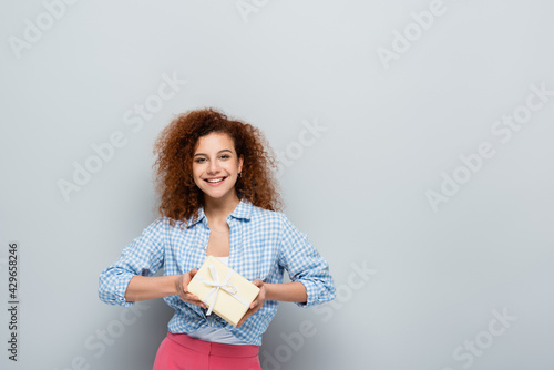 happy woman looking at camera while holding present on grey background