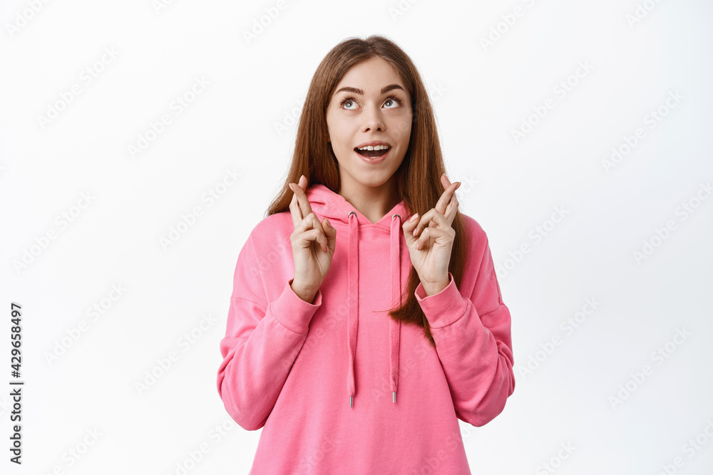 Excited girl makes wish with crossed fingers, looks aside at logo while praying, pleading for dream come true, stands against white background Excited girl makes wish with crossed fingers, looks aside at logo while praying, pleading for dream come true, stands against white background