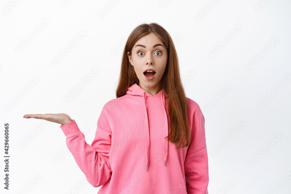 Excited young woman gasping amazed, saying wow, holding empty copy space on palm, spread hand sideways, display product, standing over white background
