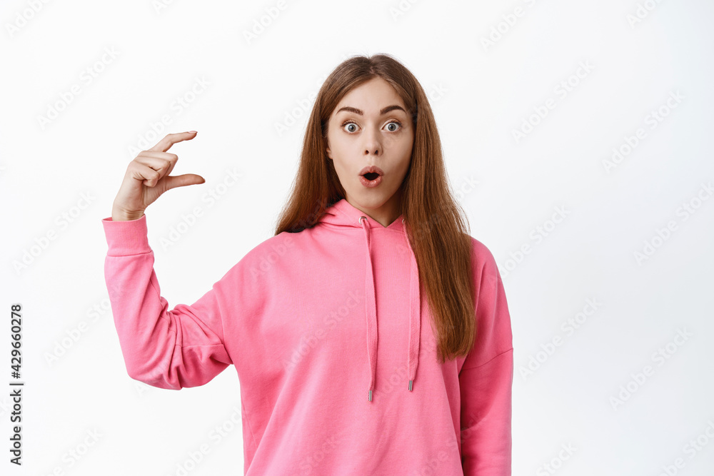 Portrait of impressed girl showing little thing, display tiny little object, looking amazed at camera, gasping and say wow, standing over white background Portrait of impressed girl showing little thing, display tiny little object, looking amazed at camera, gasping and say wow, standing over white background