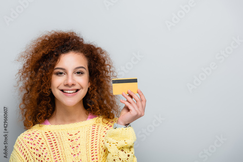 joyful woman in openwork sweater holding credit card isolated on grey