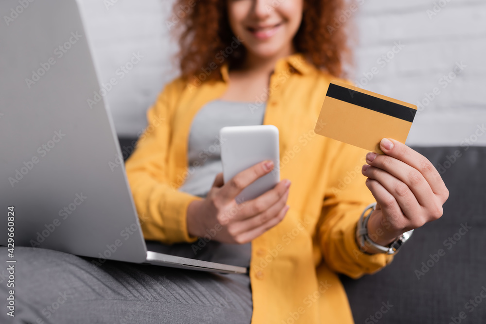 partial view of woman holding smartphone and credit card near laptop on blurred background partial view of woman holding smartphone and credit card near laptop on blurred background