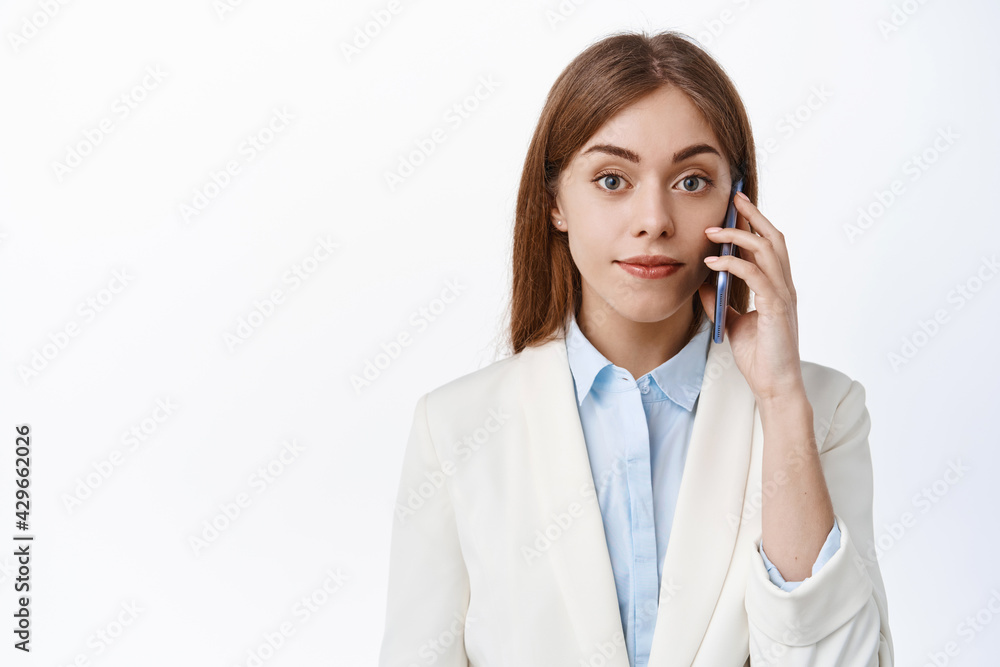 Elegant professional woman talks on mobile phone, look at camera confident and determined, making call at work, standing over white background Elegant professional woman talks on mobile phone, look at camera confident and determined, making call at work, standing over white background