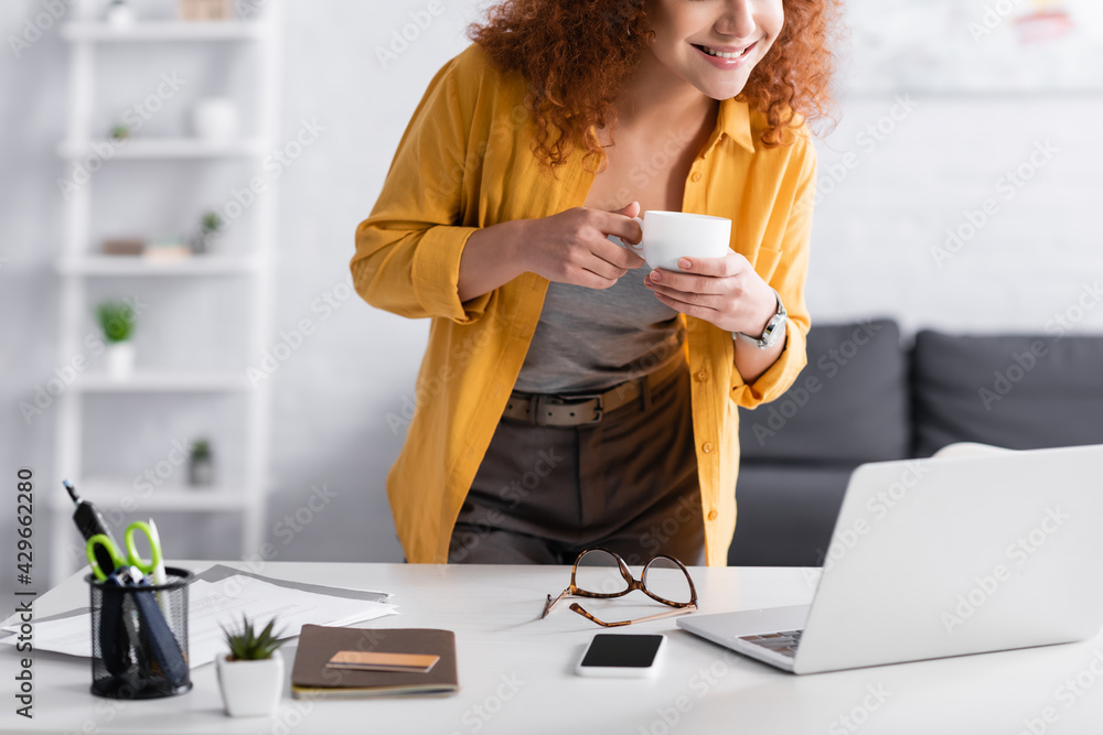 cropped view of freelancer holding coffee cup while standing near laptop cropped view of freelancer holding coffee cup while standing near laptop