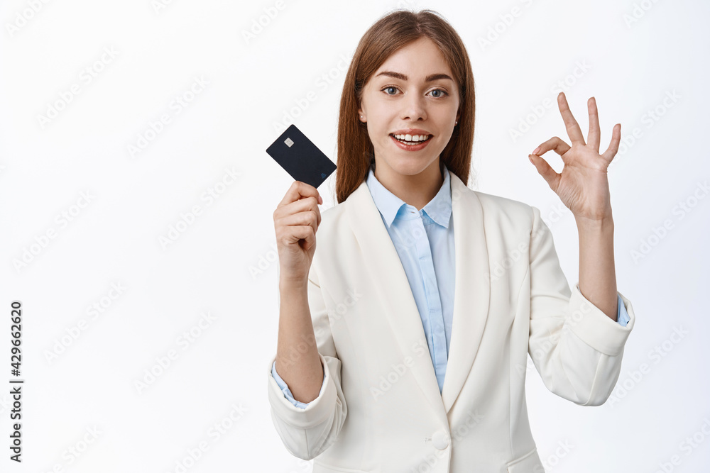 Elegant ceo, woman in business suit shows plastic credit card and okay sign, everything alright, under control, stands against white background Elegant ceo, woman in business suit shows plastic credit card and okay sign, everything alright, under control, stands against white background