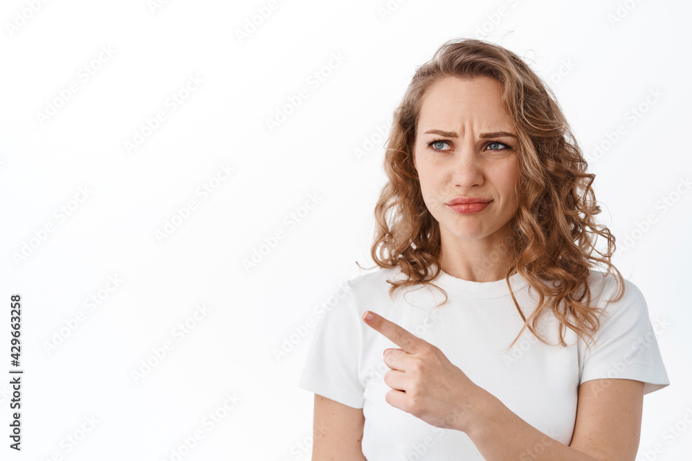 Skeptical girlfriend with blond hair, smirk and frown, looking with judgemental and displeased face at copyspace, pointing aside, standing over white background Skeptical girlfriend with blond hair, smirk and frown, looking with judgemental and displeased face at copyspace, pointing aside, standing over white background