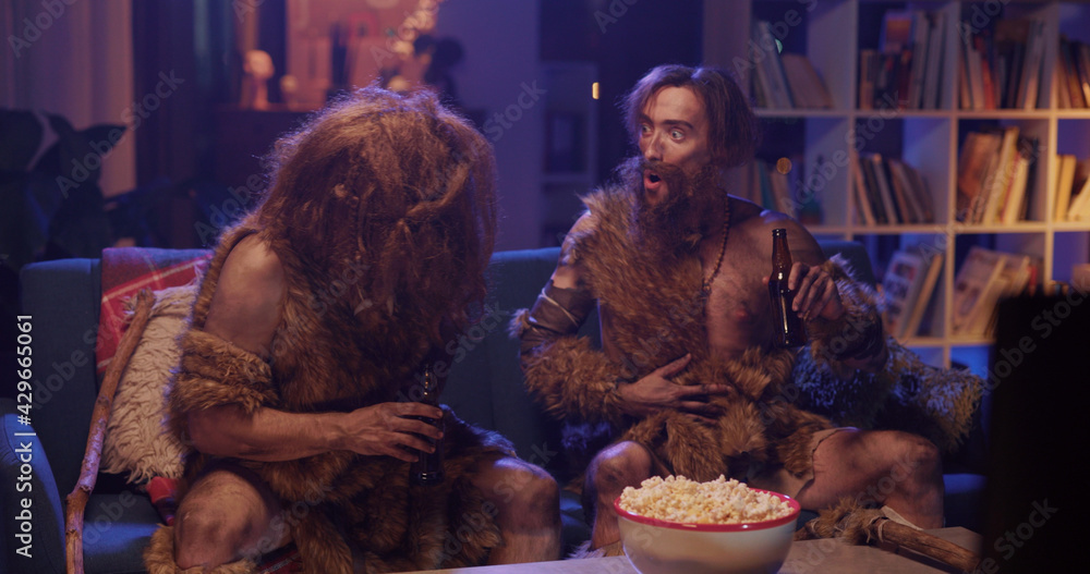 Prehistoric couple of wild people in fur clothes drinking beer throwing eating popcorn having fun in modern living room house. Entertainment. Movie concept.