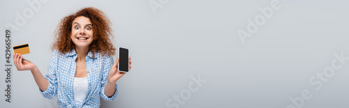 excited woman holding smartphone with blank screen and credit card isolated on grey, banner