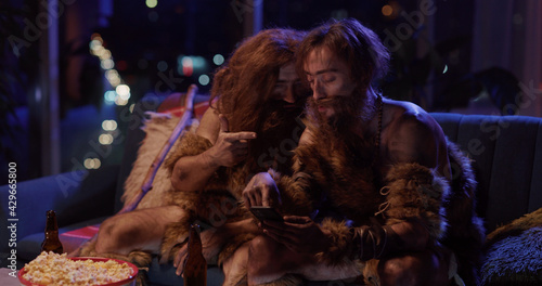 Couple of Wild Cave People Men Wearing Animal Fur Staying in Modern House Aoartment Using Phone Playing Social Media Aops Interacting Technology. © Fractal Pictures