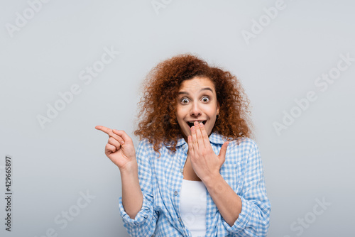 shocked woman pointing with finger and covering mouth with hand isolated on grey