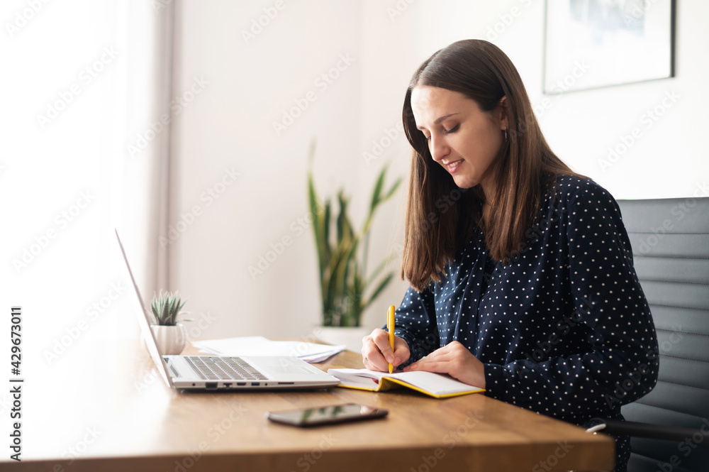 Smiling business woman writing down plans and ideas in the notepad sitting in the modern office in front of the laptop, female takes notes with a pen Smiling business woman writing down plans and ideas in the notepad sitting in the modern office in front of the laptop, female takes notes with a pen
