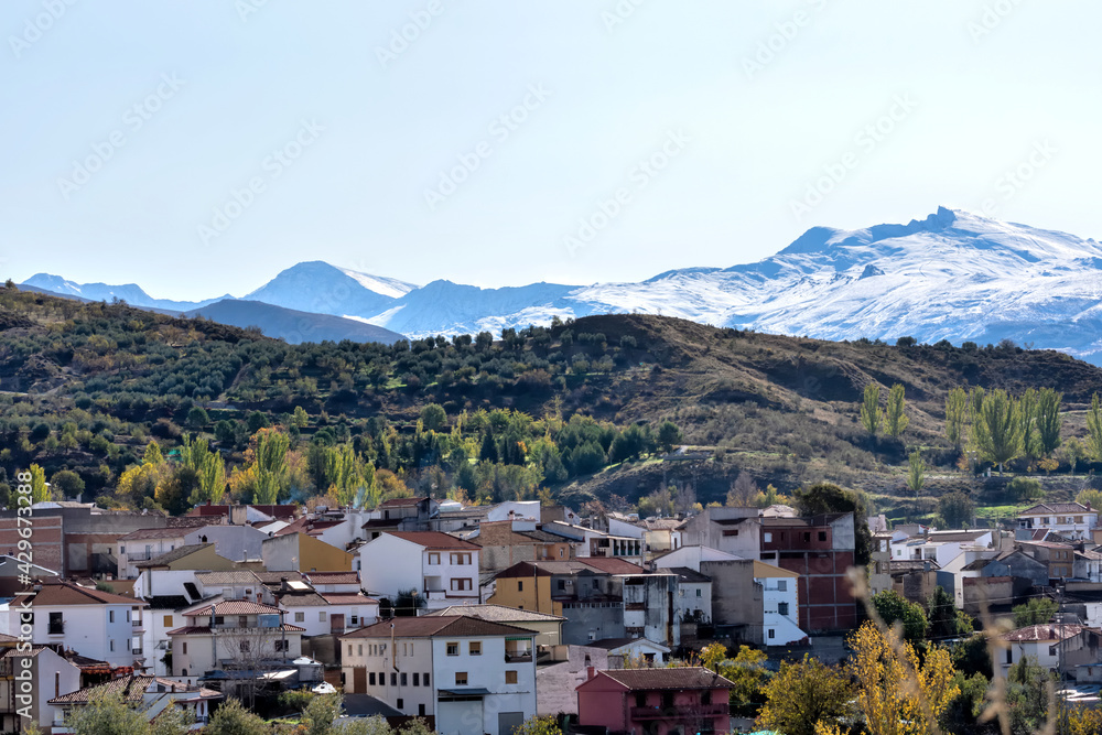 View of the Andalusian town Beas de Granada among trees with the high snowy peaks of Sierra Nevada in the background View of the Andalusian town Beas de Granada among trees with the high snowy peaks of Sierra Nevada in the background