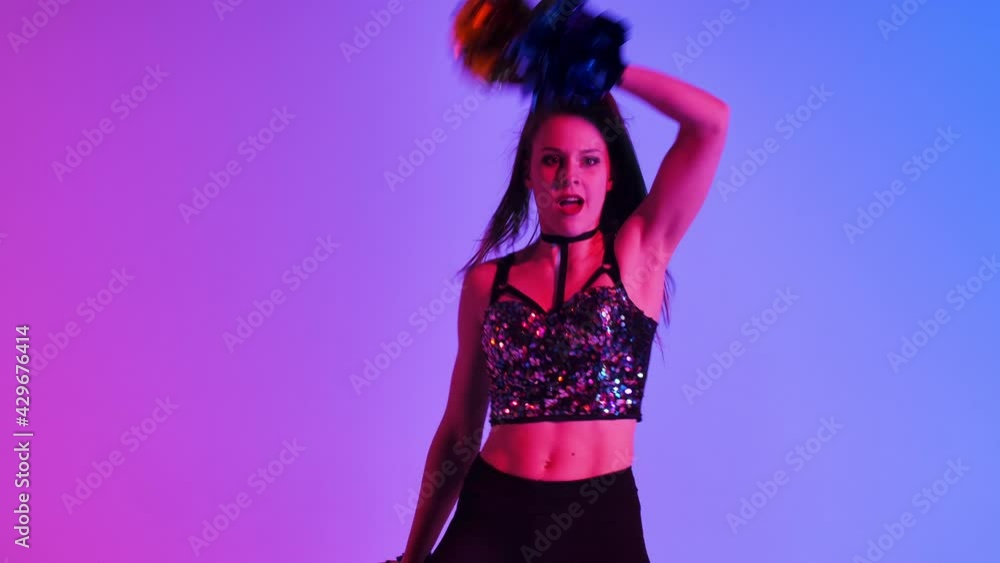 Energetic cheering dance performed by cute cheerleader with pompoms in hands against blue pink studio background. Adorable female moves hands and hips, whirls and waves hair. Close up. Slow motion. Energetic cheering dance performed by cute cheerleader with pompoms in hands against blue pink studio background. Adorable female moves hands and hips, whirls and waves hair. Close up. Slow motion.