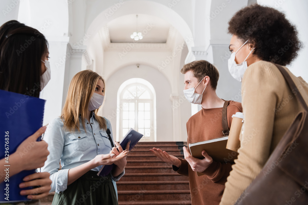 Students in medical masks pointing with hands near multiethnic friends with notebook and coffee Students in medical masks pointing with hands near multiethnic friends with notebook and coffee