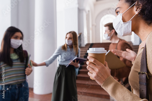 African american student in medical mask holding coffee to go near friends on blurred background