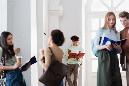 Smiling african american student looking at notebook near blurred people in university