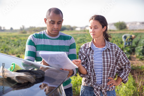 Focused couple of farmers standing outdoors near car on background with vegetable plantation, discussing documents..
