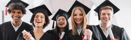 Φωτογραφία Multiethnic students in caps holding diplomas and smiling at camera, banner