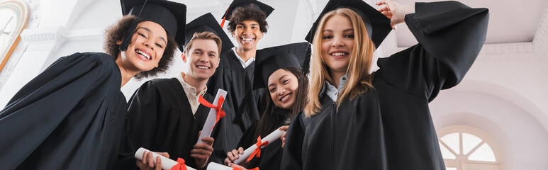 Low angle view of smiling interracial graduates holding diplomas, banner