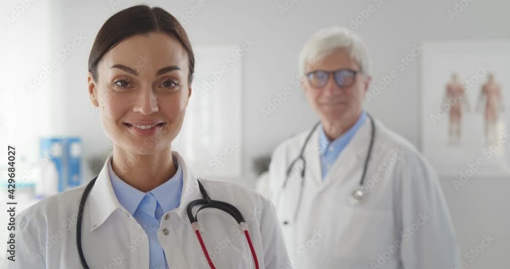 Portrait of young woman doctor smiling at camera with aged colleague standing on background