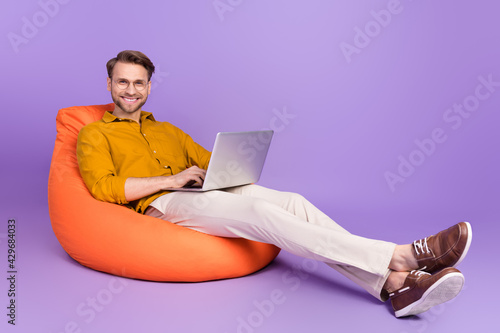 Full length body size photo of guy sitting in beanbag using laptop isolated on vivid purple color background