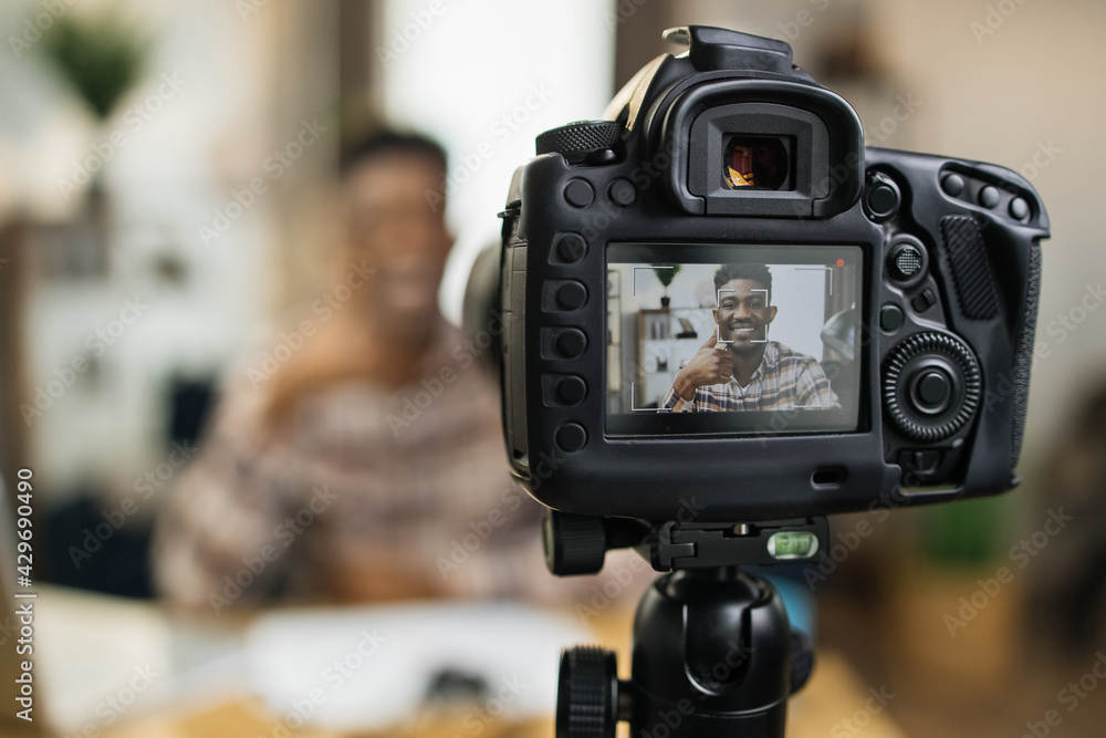 Blur background of smiling black man sitting at table with wireless laptop and recording video. Focus of modern professional camera. Online tutorial concept.