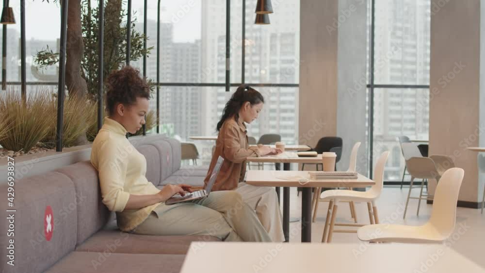 Medium long side view of two young multiethnic women sitting on couch at restaurant tables, working, keeping safe social distance Medium long side view of two young multiethnic women sitting on couch at restaurant tables, working, keeping safe social distance