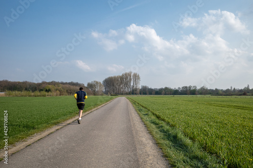 Outdoor sunny view of runner jog on small road in suburb area surrounded with agricultural field with green meadow in spiring season against blue sky.