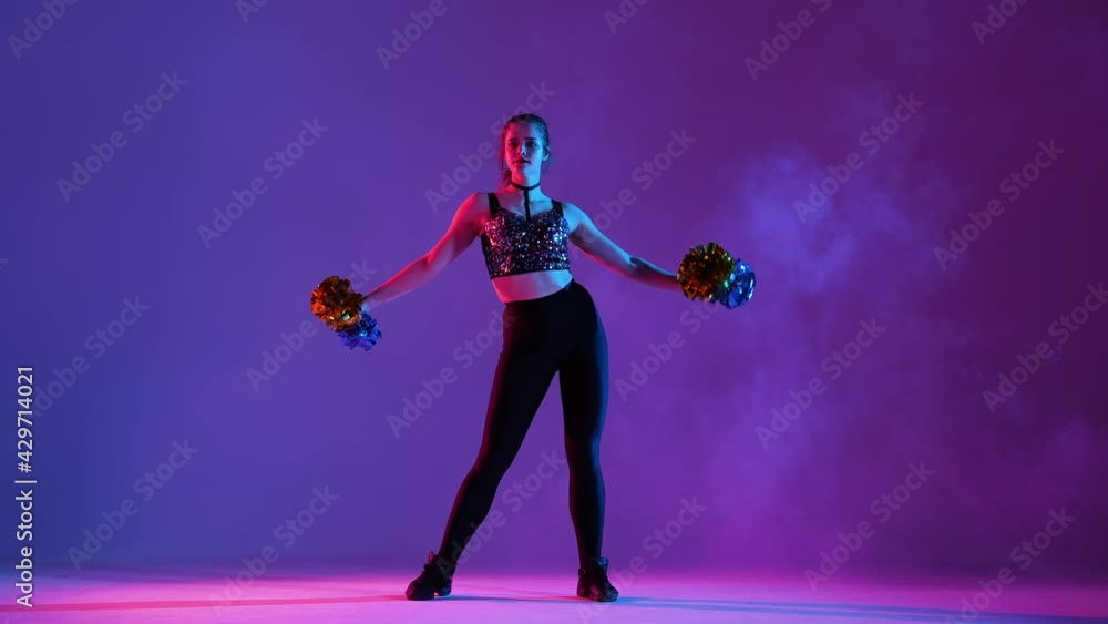 Young woman with an athletic slim body is practicing cheerleading, dancing with pompoms on a purple studio background with smoke. Cheerleader waves hands and sensually moves her hips. Slow motion. Young woman with an athletic slim body is practicing cheerleading, dancing with pompoms on a purple studio background with smoke. Cheerleader waves hands and sensually moves her hips. Slow motion.