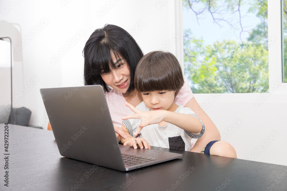 Asian woman with a child works on computer. Concept of work from home and home family education. Mom and son are working on a laptop at home.