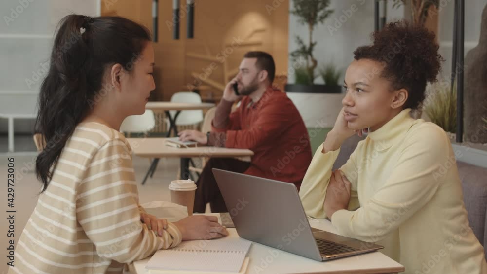 Side view medium of two multiethnic girlfriends sitting at restaurant table with notebooks and portable computer on, smiling and chatting, blurred man talking on phone on background Side view medium of two multiethnic girlfriends sitting at restaurant table with notebooks and portable computer on, smiling and chatting, blurred man talking on phone on background