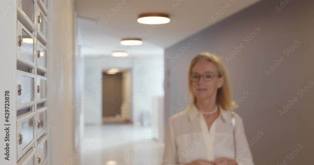 Senior female in eyewear opening postbox standing in hallway and holding envelope with invitation. Senior female in eyewear opening postbox standing in hallway and holding envelope with invitation.