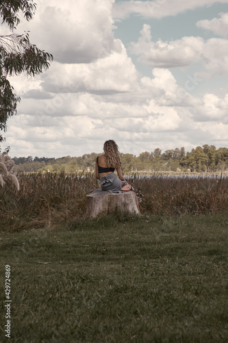 mujer rubia de cabello rizado sentada de espaldas a la camara mirando el horizonte en un paisaje de campo