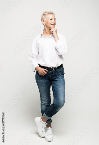 Photographie Full length photo of stylish elderly woman wearing jeans and white blouse with short white hair posing on a light gray background