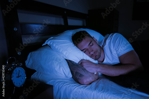 Handsome man asleep in bed at night with traditional alarm clock on the bedside table next to him. 