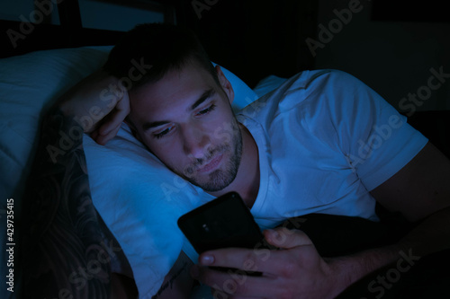 Handsome man looking at his mobile phone in bed at night with glow of phone lighting his face