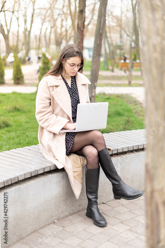 Vertical full length photo of a modern businesswoman working on a laptop in the park siting on the bench