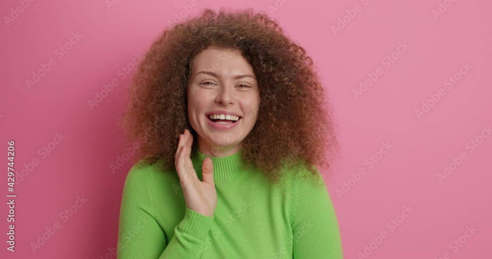 Positive beautiful young Caucasian woman smiles happily winks eye has carefree expression feels glad wears casual green poloneck isolated over pink background. People emotions happiness concept Positive beautiful young Caucasian woman smiles happily winks eye has carefree expression feels glad wears casual green poloneck isolated over pink background. People emotions happiness concept