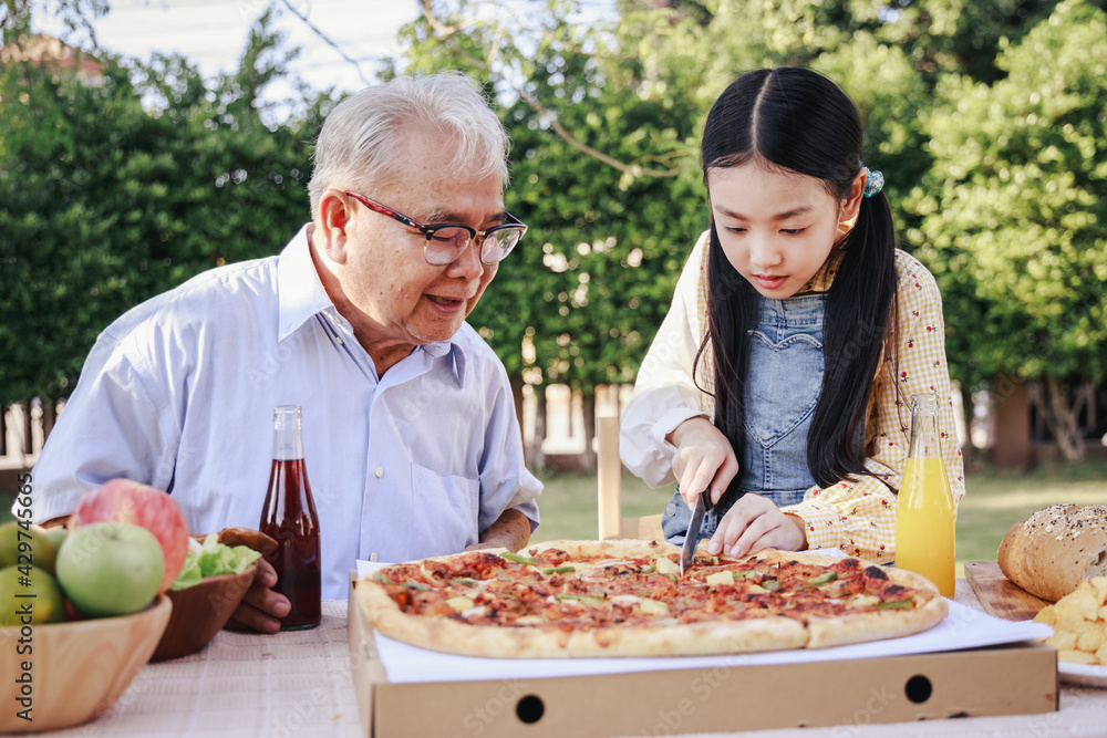 Granddaughter enjoying slicing pizza for retirement grandfather in home garden. Happy senior life after retirement with family concept.