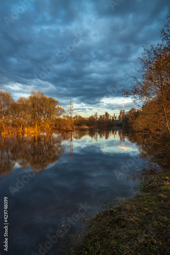 Rural landscape with a river.Trees, bushes and the ground are illuminated by the rays of the setting sun.The sky is covered with clouds.Lead clouds are reflected in the water.A center on the horizon