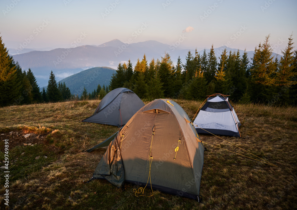 Tourist camp with three tents set up on meadow near forest. Morning sunlight on tent and surrounding nature. Silhouettes of high mountains on background.