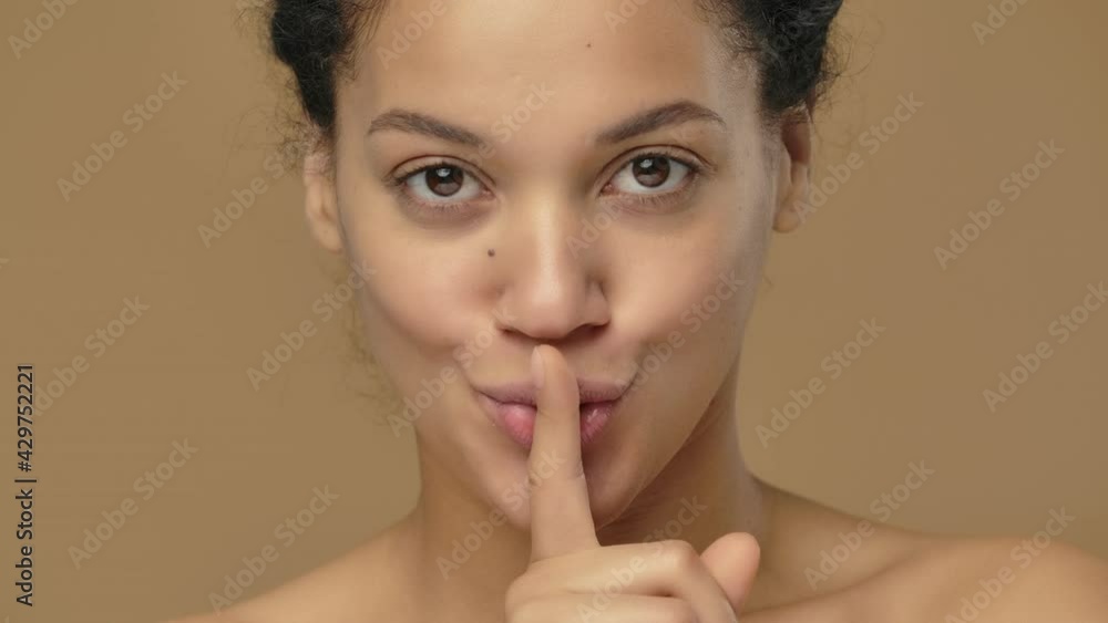 Beauty portrait of young African American woman coquettishly looking at camera and showing hush gesture, secret. Black female on brown studio background. Close up. Slow motion ready, 4K at 59.94fps. Beauty portrait of young African American woman coquettishly looking at camera and showing hush gesture, secret. Black female on brown studio background. Close up. Slow motion ready, 4K at 59.94fps.