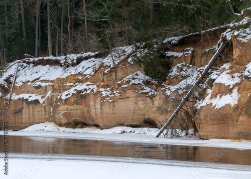 landscape with frozen river Gauja near Gudu cliffs, view of sandstone cliff from the opposite bank of the river, beautiful red rock wall, bare trees on the river bank, winter, Ligatne, Latvia photo