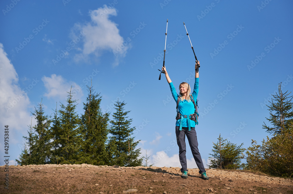 Happy blonde triumphantly raising her hands holding trekking sticks after she climbed on stone top of hill, on the background of several green trees and beautiful blue sky.