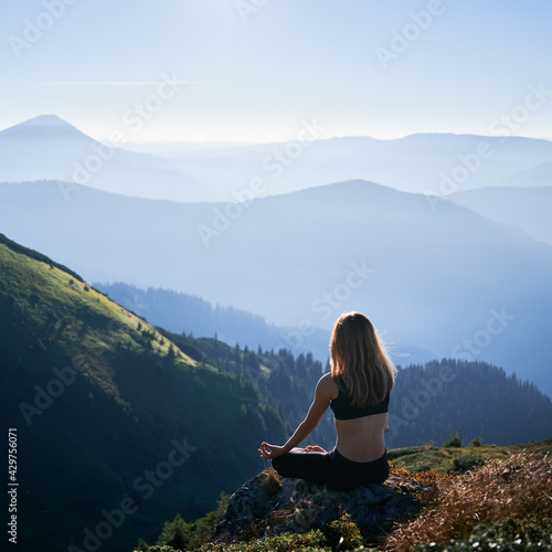Back view of brunette practicing yoga in Lotus pose in the morning. Meditating female is relaxing in the mountains. Concept of fit body.